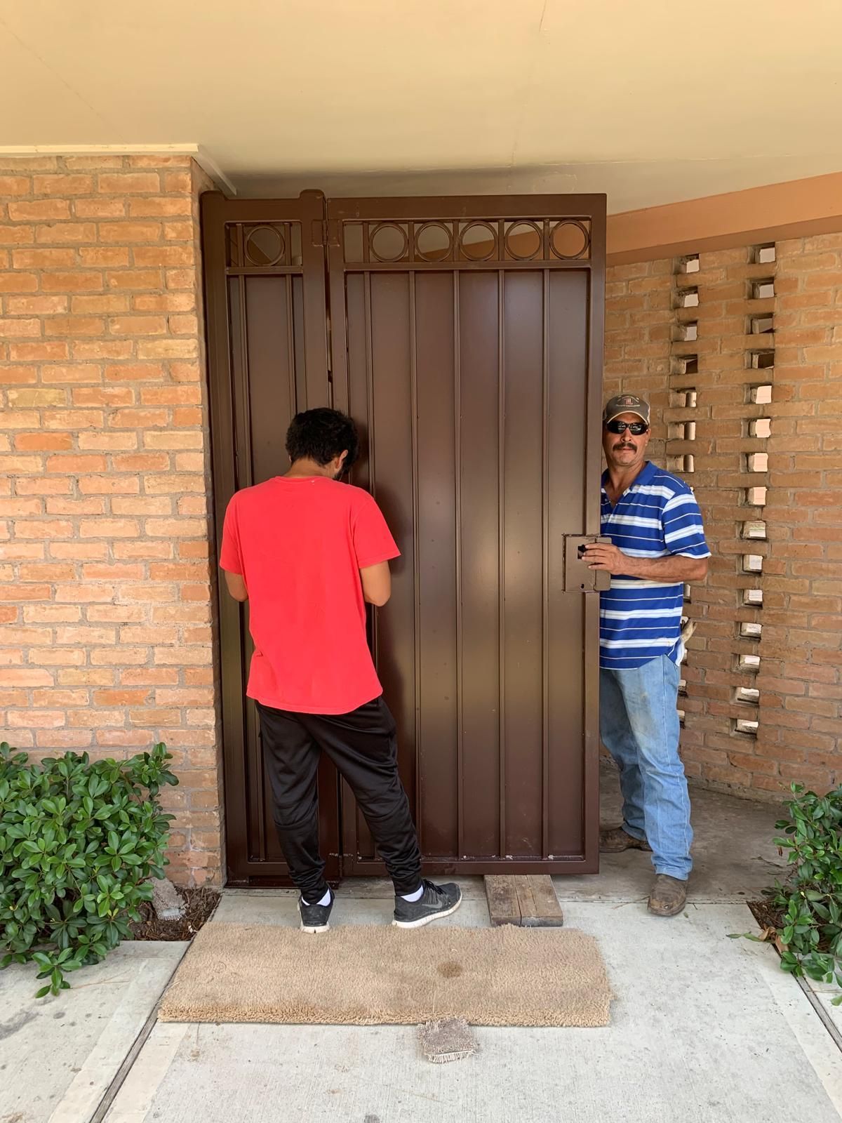 Two men installing a brown metal gate in front of a brick building.
