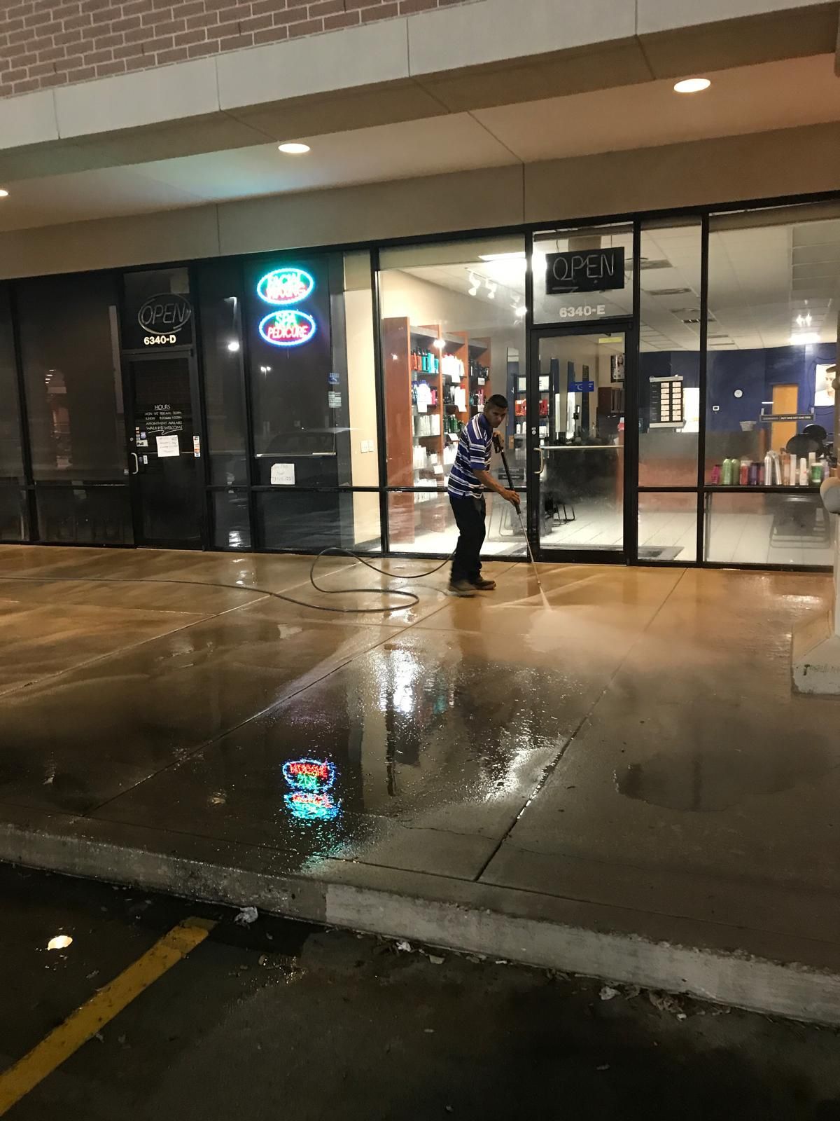 Man power washing a wet sidewalk in front of a store at night.