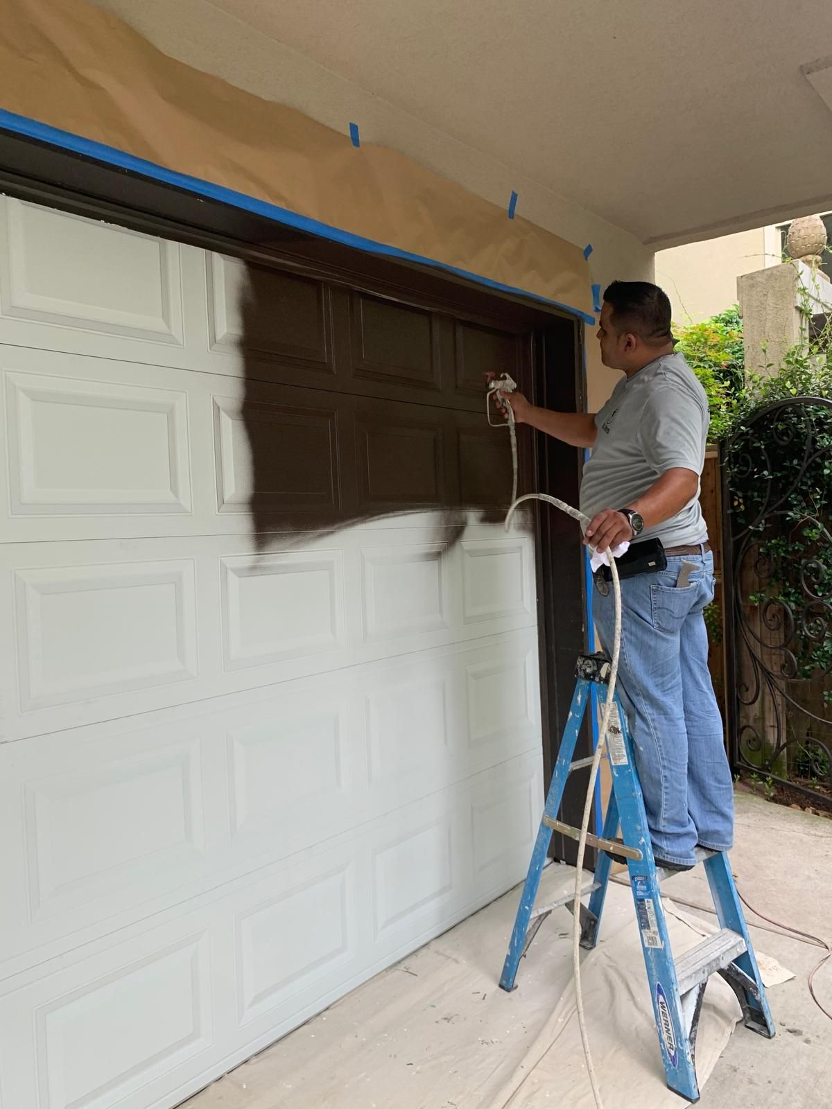 Man sprays brown paint on a white garage door from a blue ladder, exterior setting.