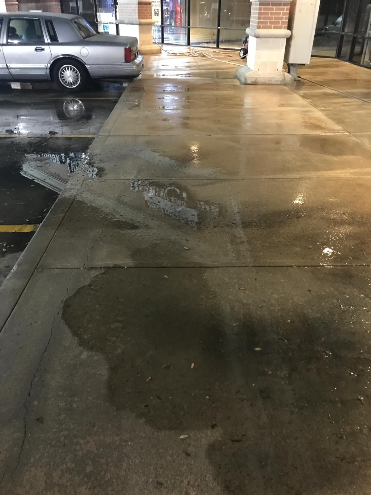 Wet concrete sidewalk with puddles and a parked car; night.