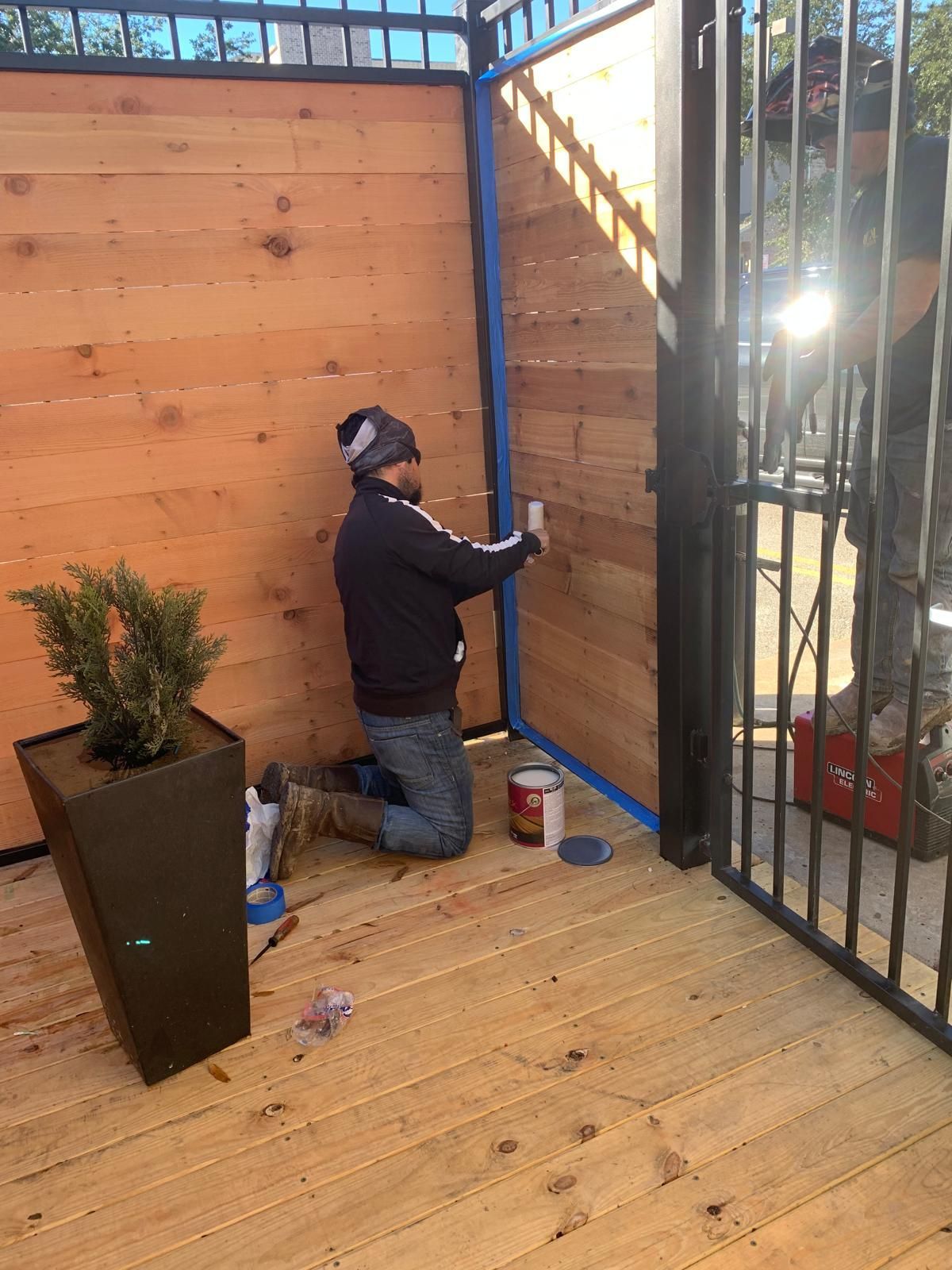 Man painting wooden fence outdoors, kneeling with tape, paint, and potted plant nearby.