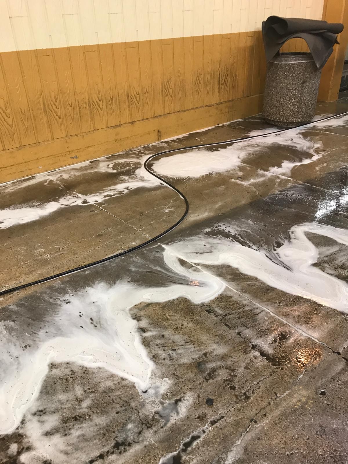 Wet concrete floor with white suds, black hose, and a trash can next to a yellow wall.