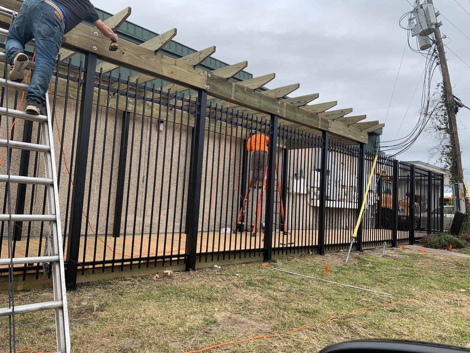 Man on ladder installing black metal fence with wooden pergola.