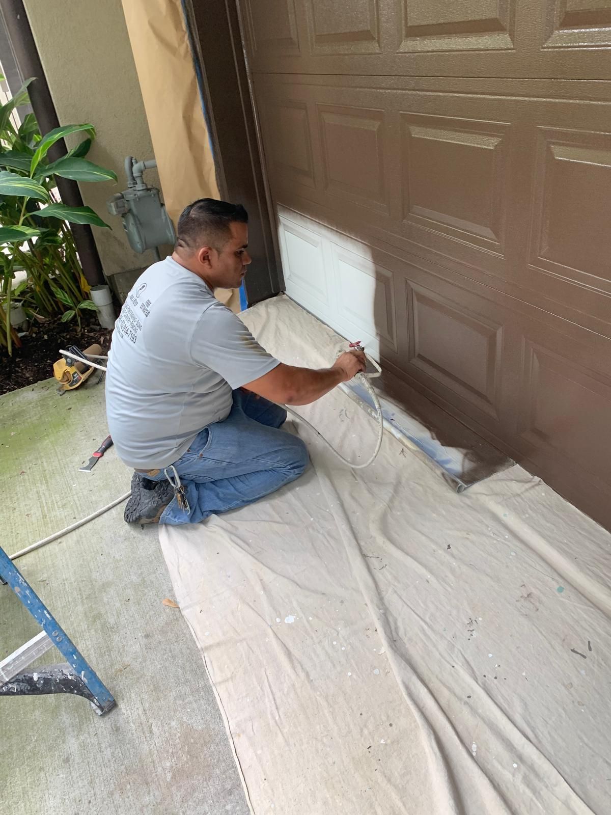 Man painting brown garage door white outdoors, kneeling on drop cloth.