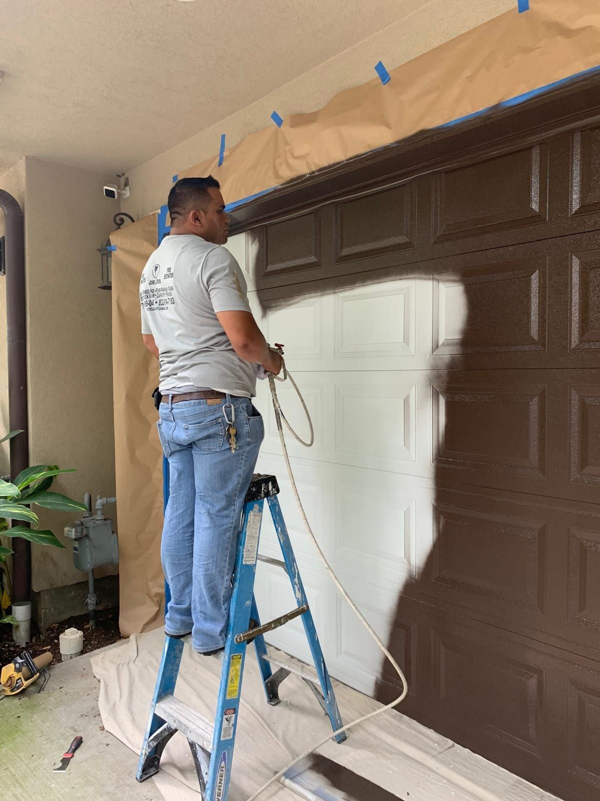 A man on a ladder sprays white paint on a brown garage door; a brown door frame is taped.