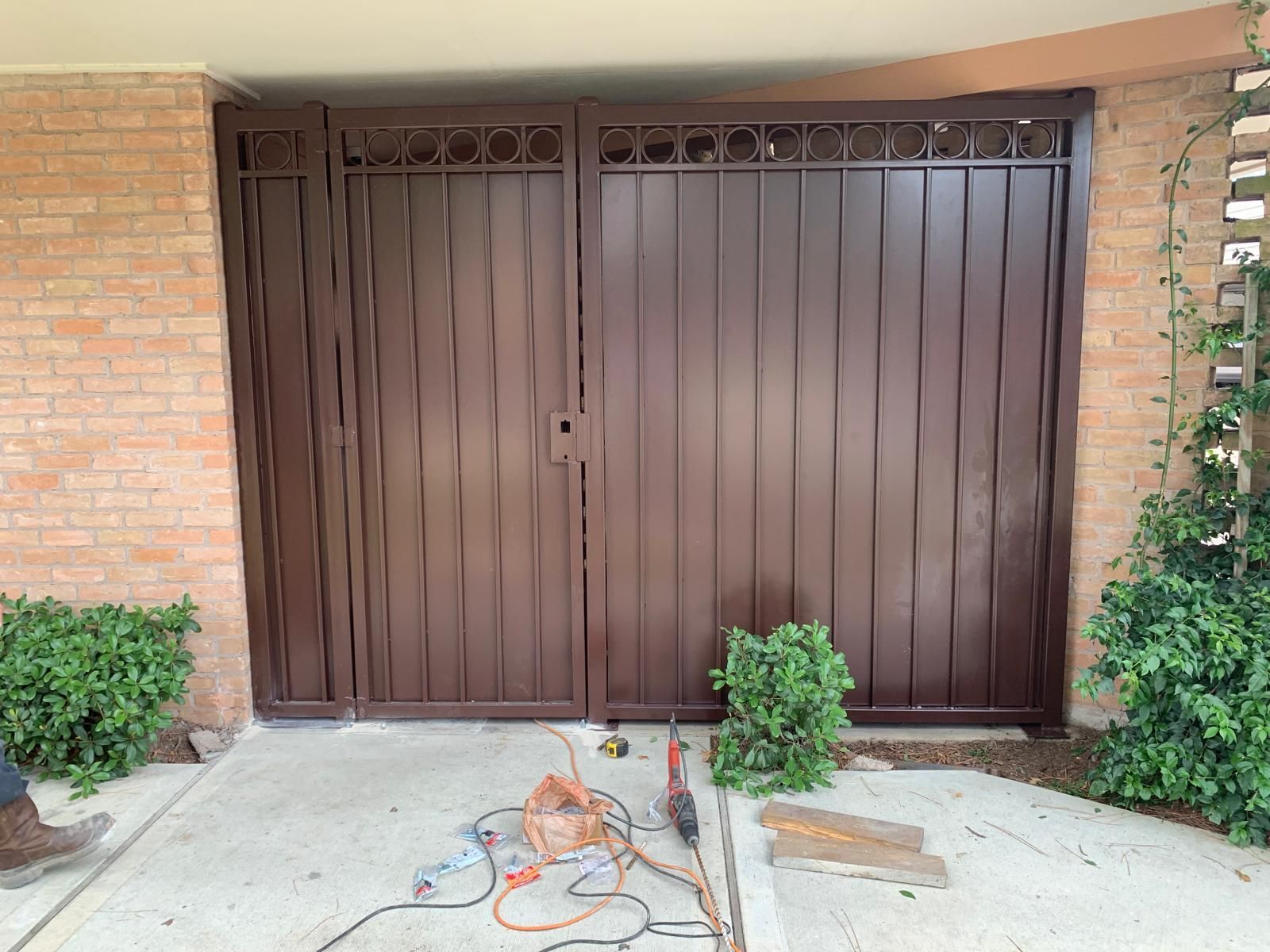 Brown metal gate in a brick building entrance with tools in front and plants on the sides.
