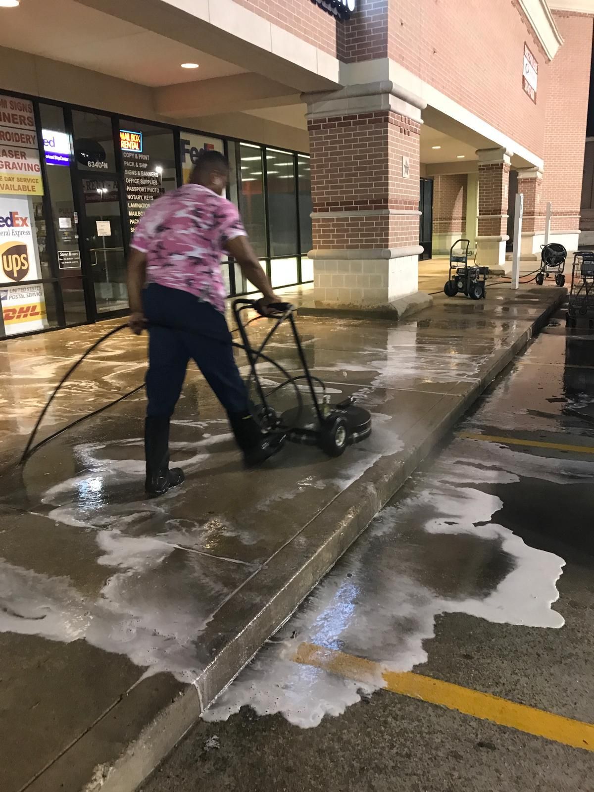 Person pressure washing a concrete sidewalk in front of a brick building with store fronts.