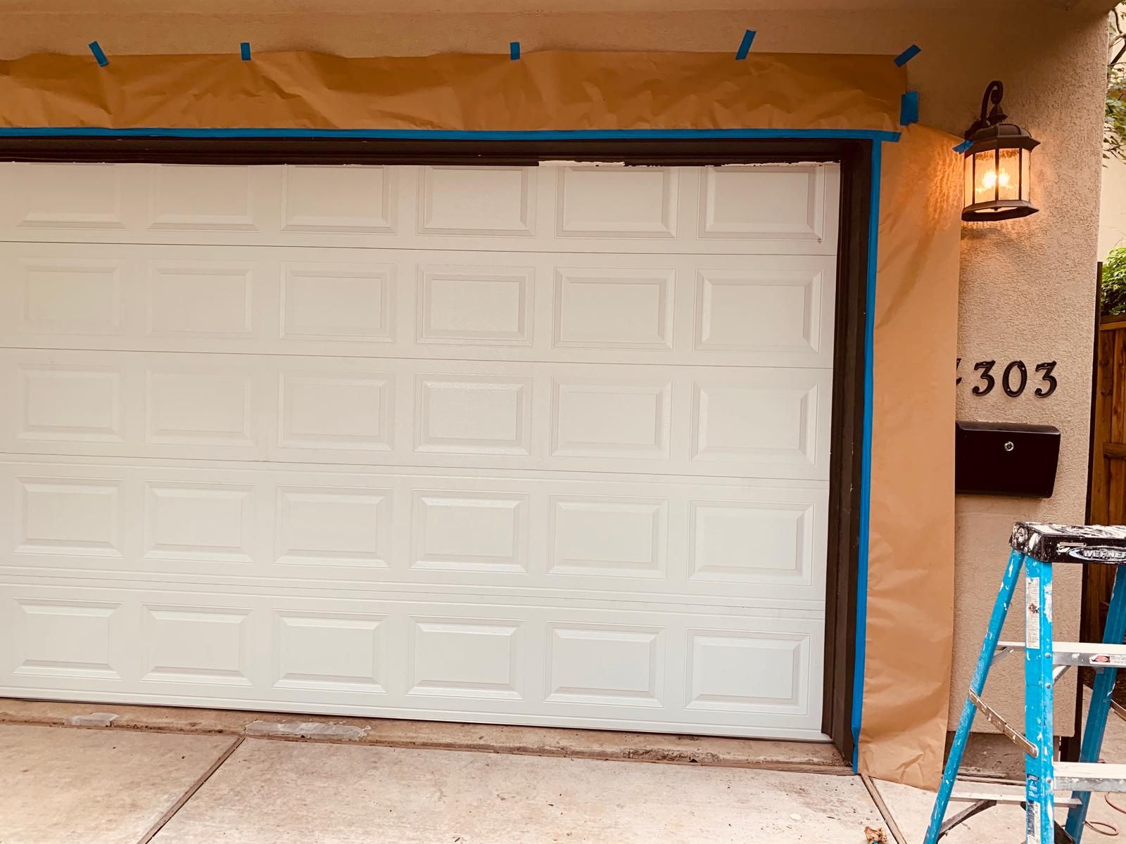 Garage door with masked trim; light on exterior wall next to house number 303; a ladder leans against the wall.