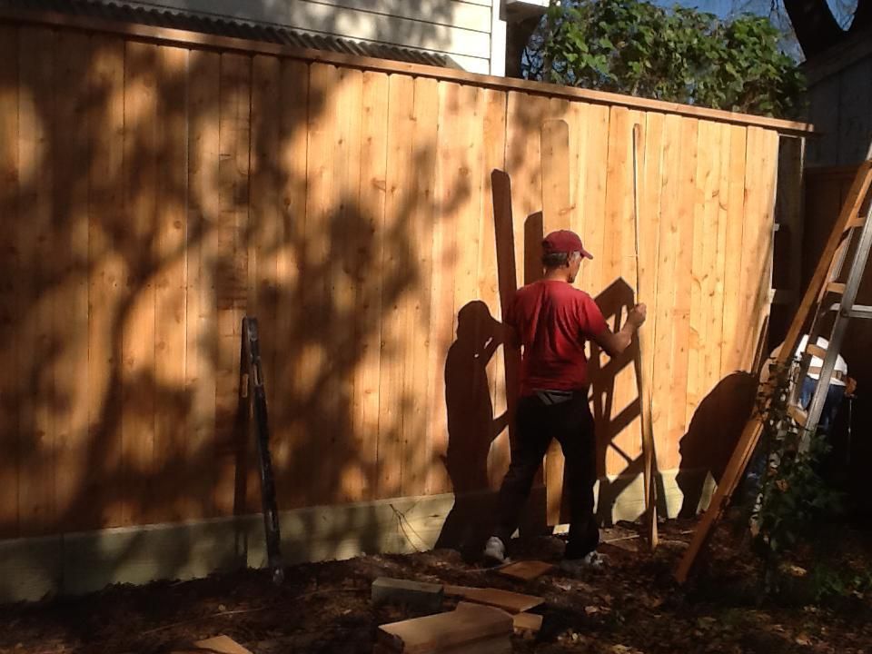 Man in red shirt building a wooden fence outdoors on a sunny day.