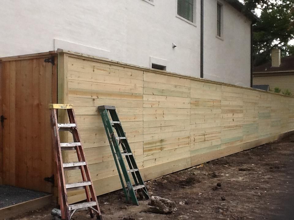 Wooden fence under construction next to a light-colored building. Two ladders lean against the fence.