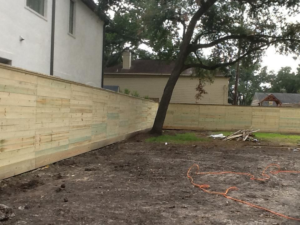 A wooden fence surrounds a vacant lot with a tree; a house is on the left and a house is in the background.