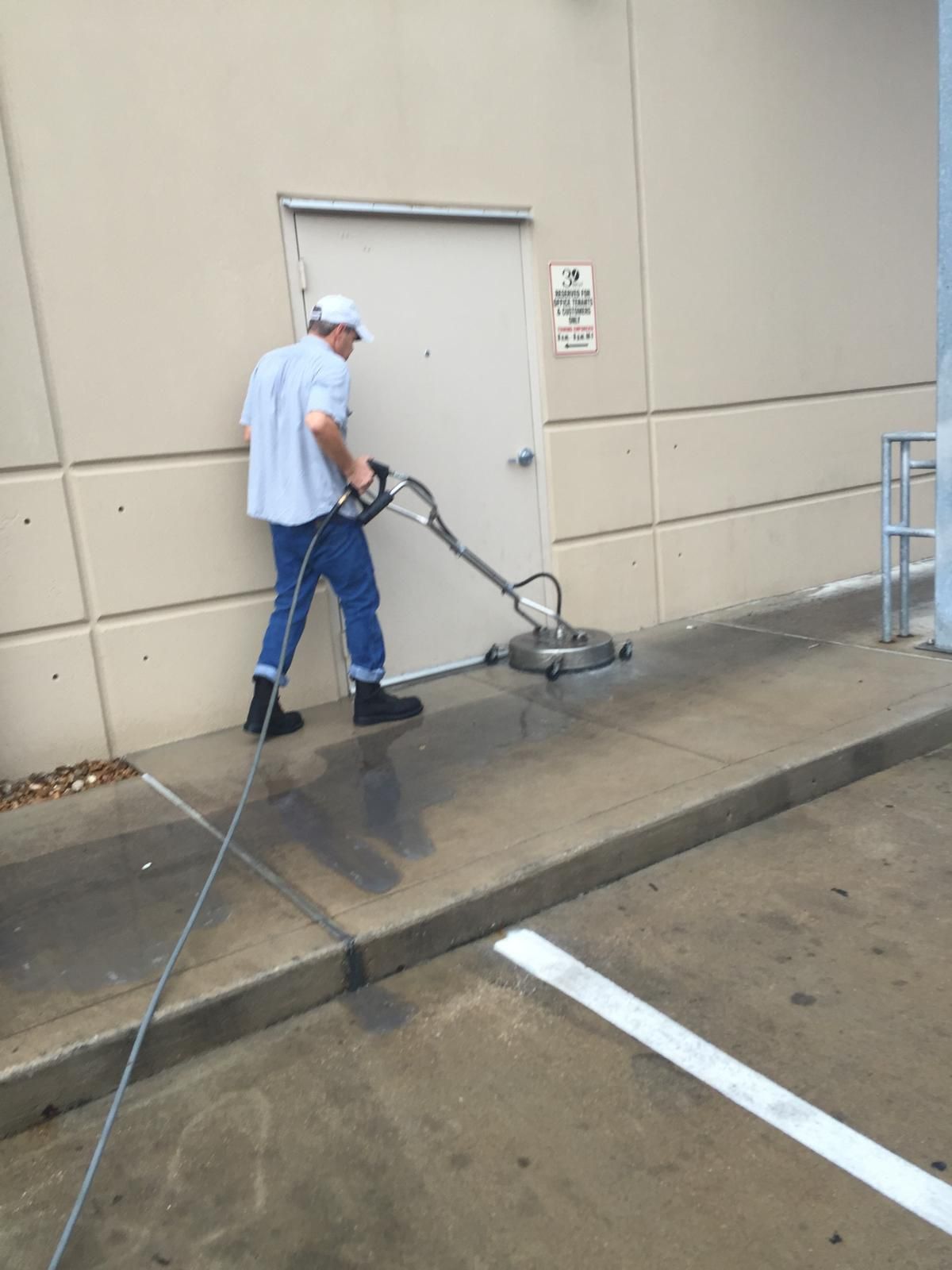 Man power washing a concrete sidewalk in front of a building with a gray door.
