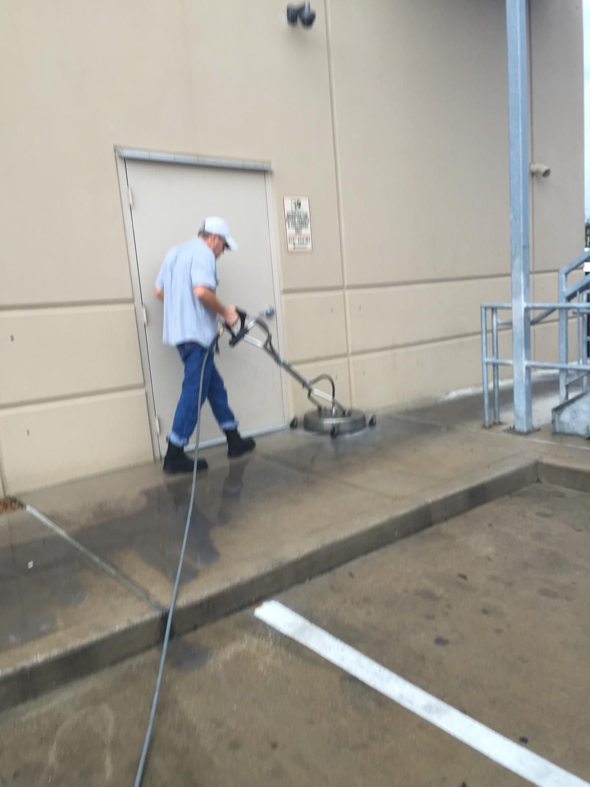 Man power washing a concrete walkway next to a building with a metal door and a parking line.