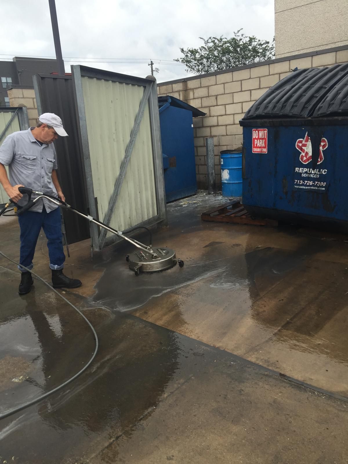 Man power washing a concrete surface near a dumpster and a building.