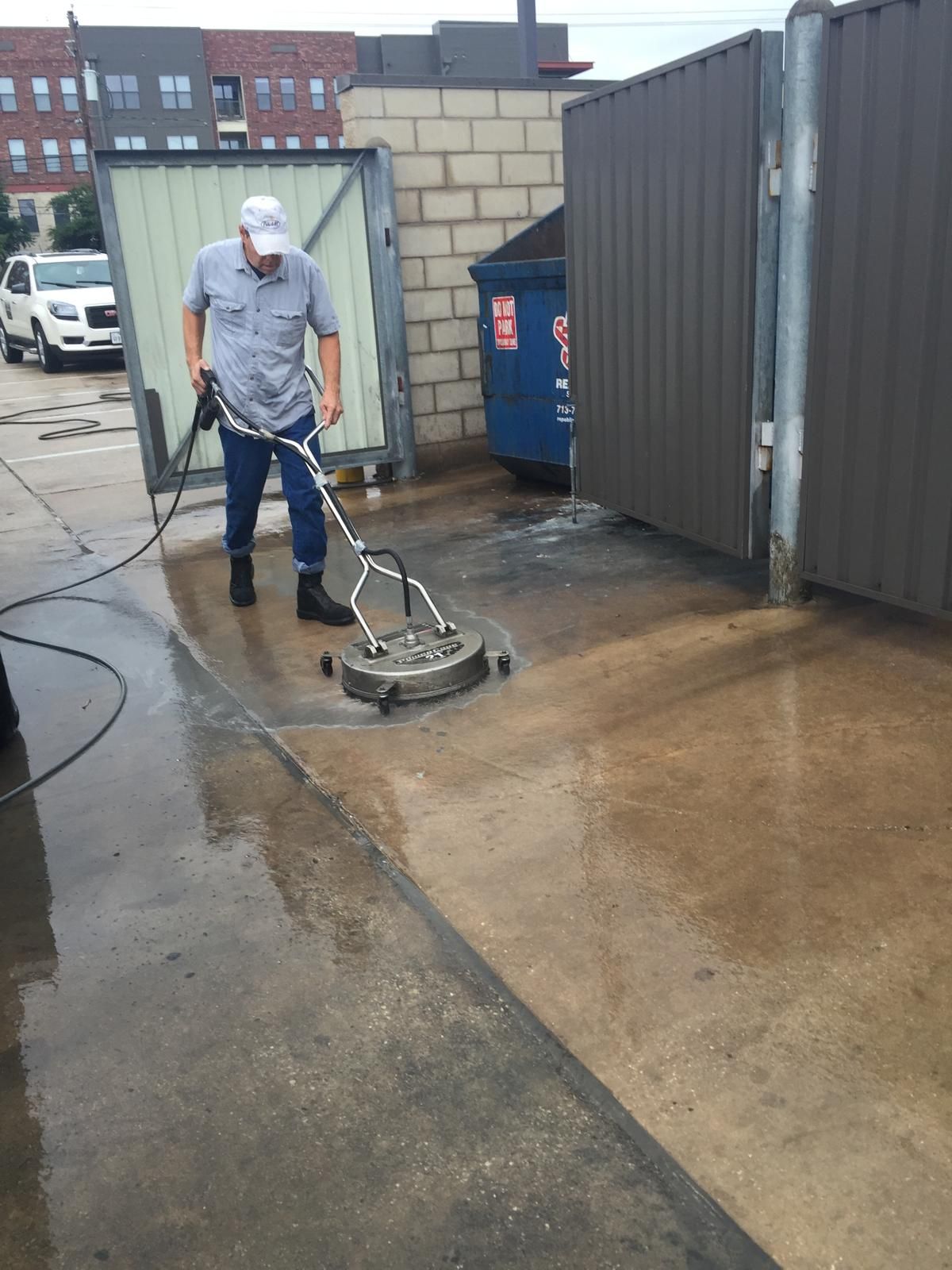 Man pressure washing a concrete area next to metal fencing and a dumpster.