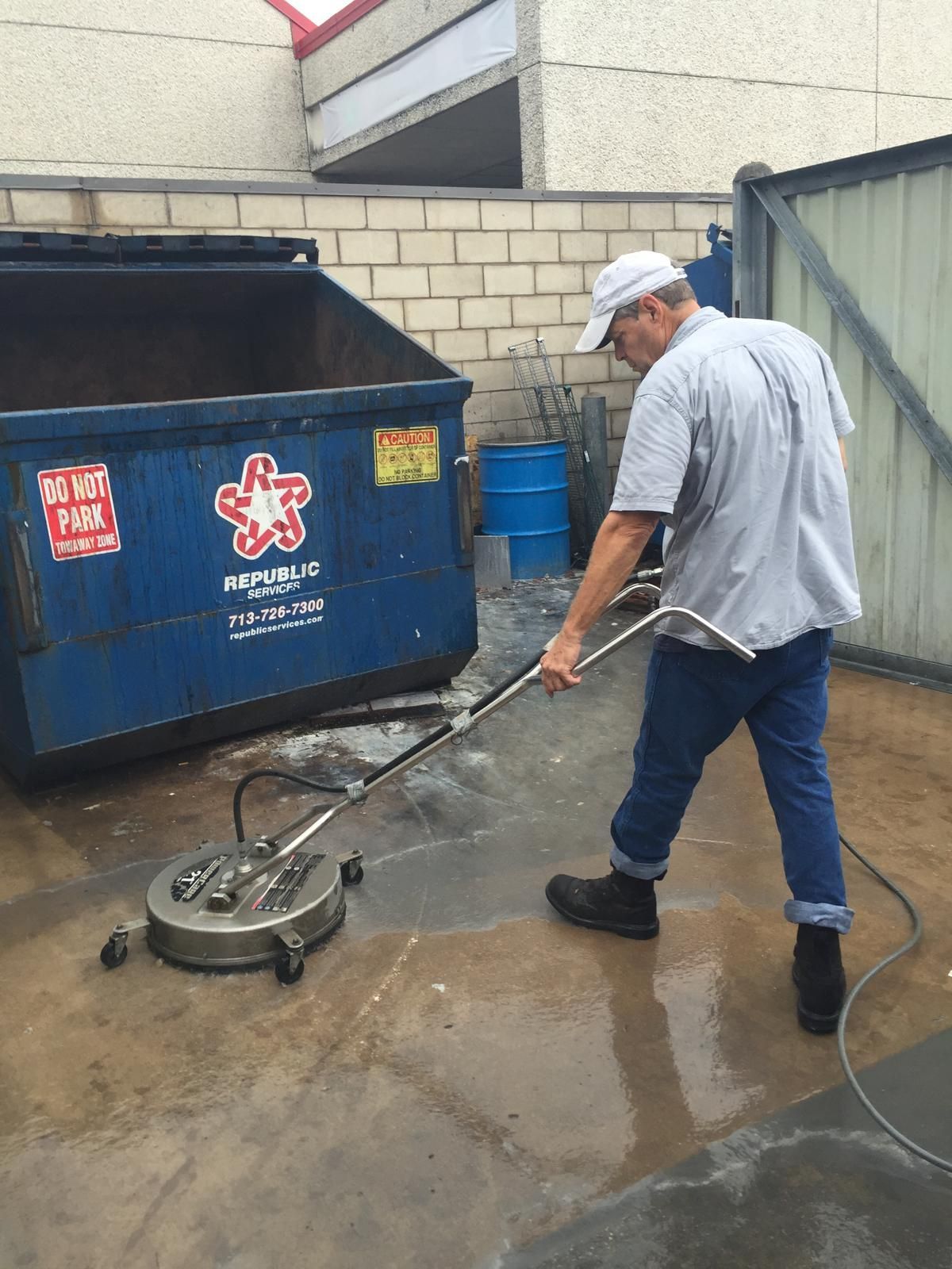 Man pressure washing concrete with a circular cleaner near a dumpster.