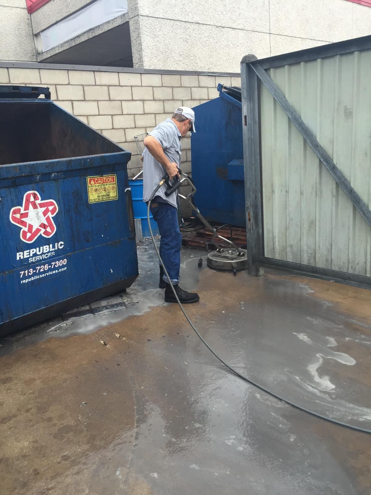 Man power washing a concrete area with a dumpster and metal gate nearby.