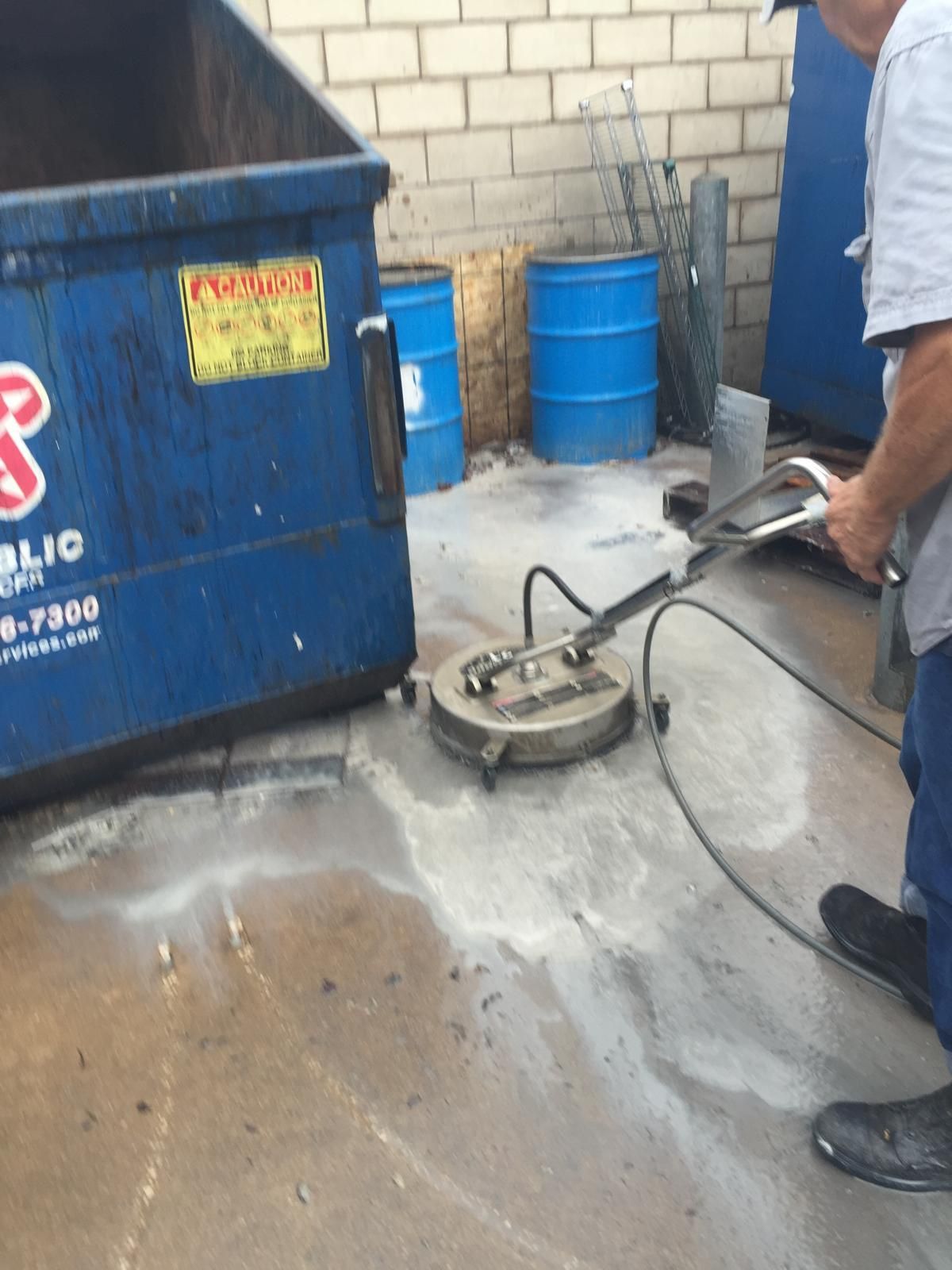 A person using a pressure washer to clean a concrete surface next to a blue dumpster.