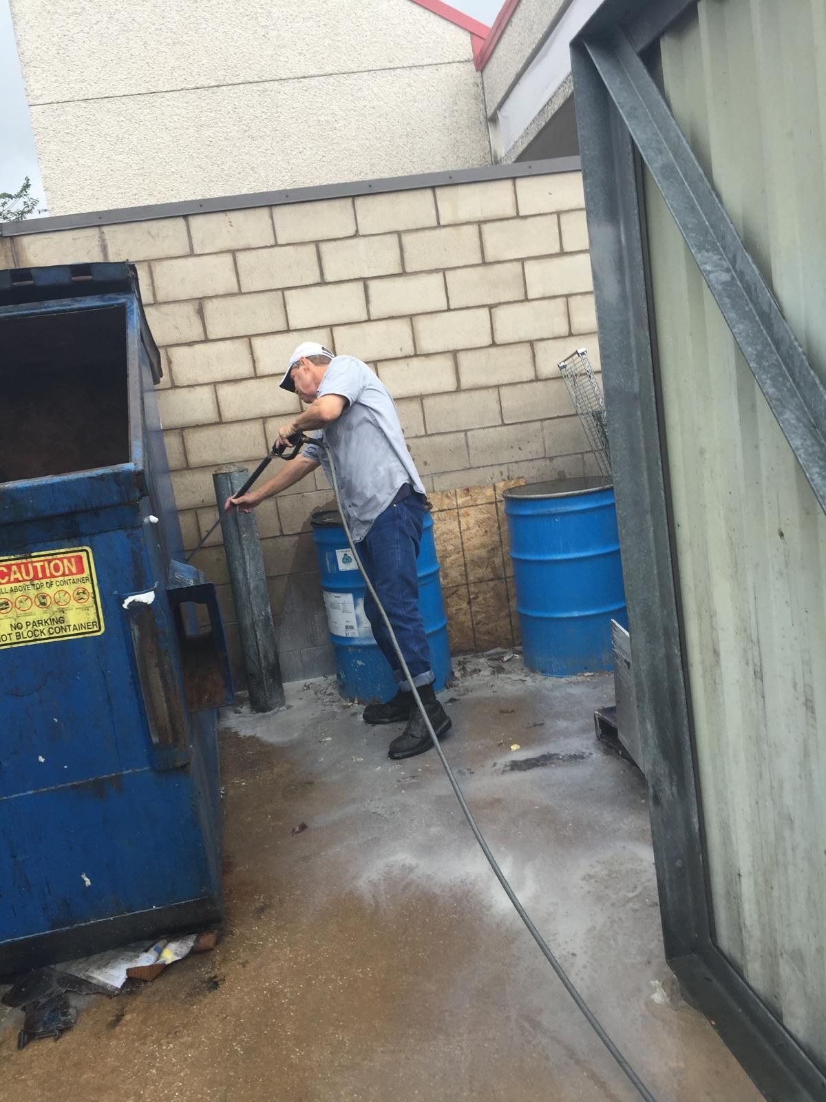 Man power washing a dumpster in an alley, wearing a white hat and gray shirt.