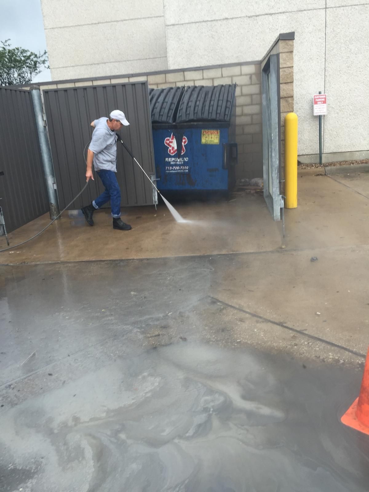 Person power washing a wet concrete area near a dumpster enclosure; exterior setting.