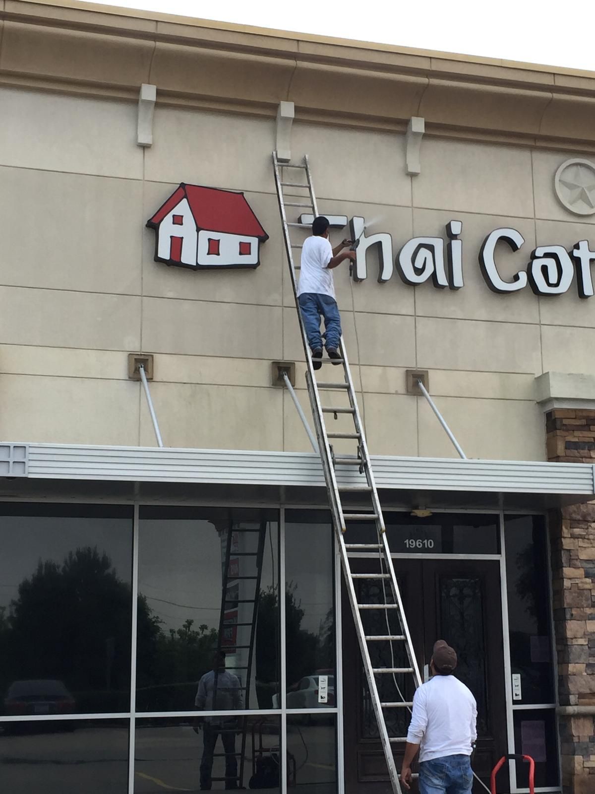 Man on ladder installing sign above a Thai Cottage restaurant entrance. Another man watches from below.