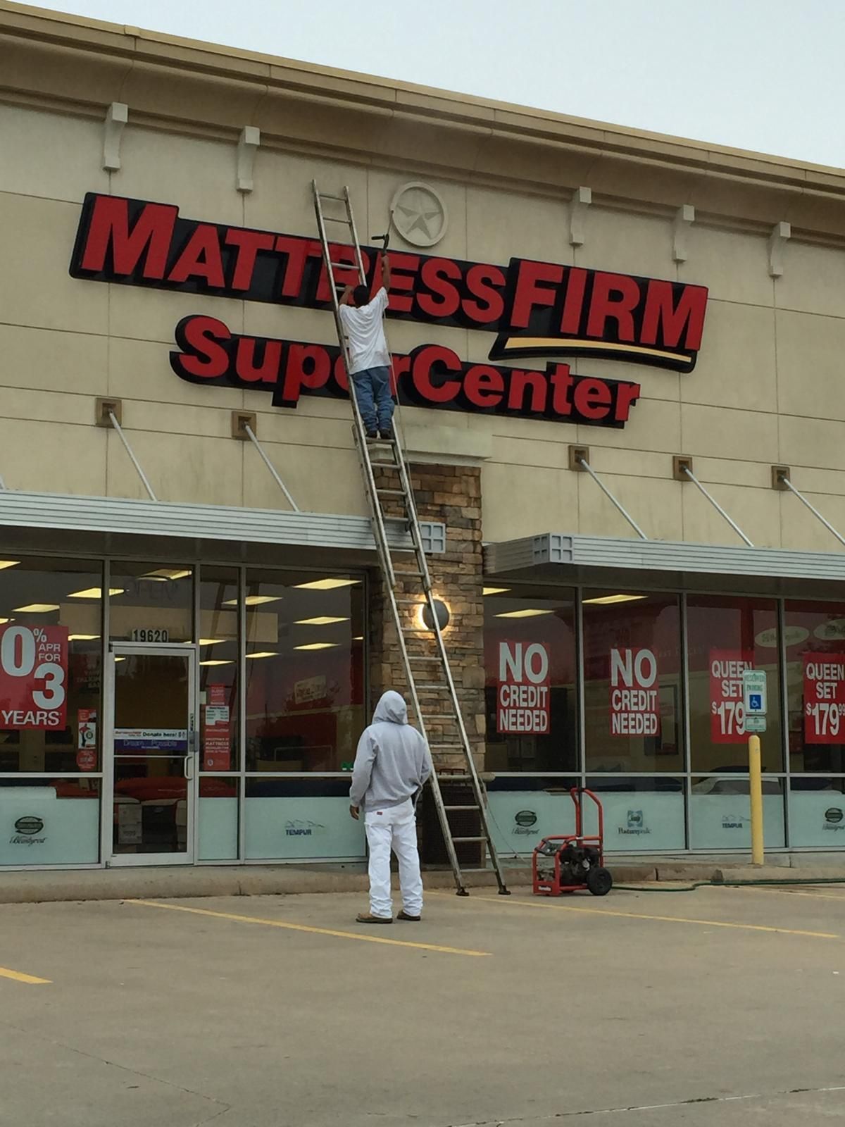 Two people painting a Mattress Firm sign on a commercial building. One is on a tall ladder, the other stands below.