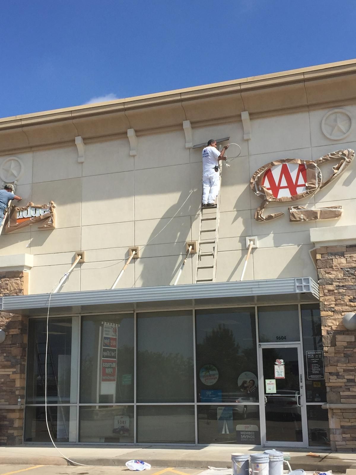 Workers painting exterior of a commercial building, standing on ladders. Building has awning, beige walls, and a partially removed sign.