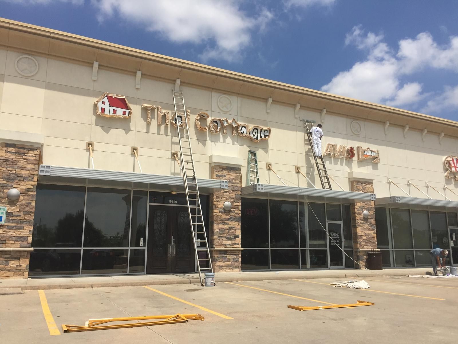 Exterior of a strip mall; workers on ladders painting signage; sunny day.