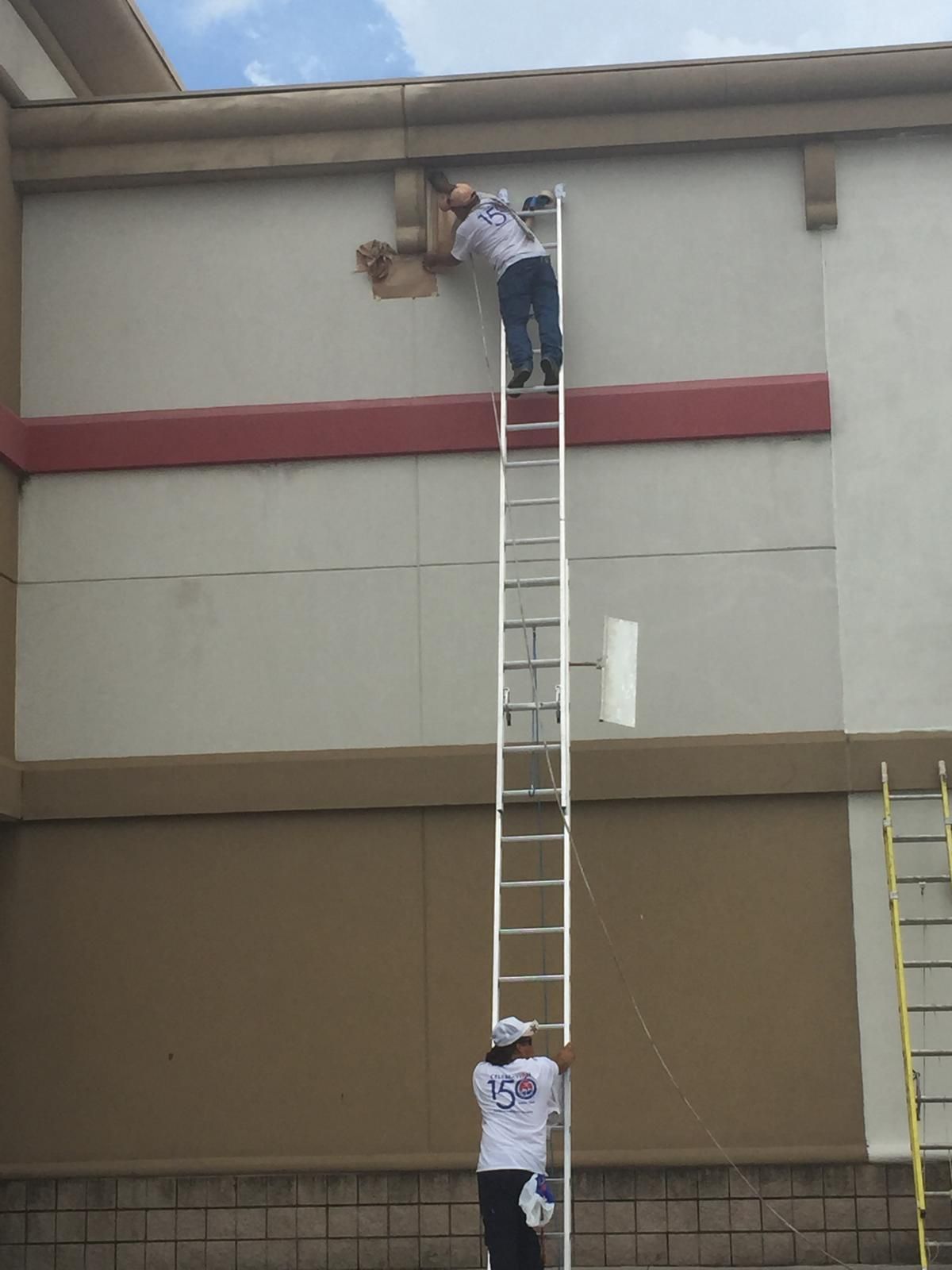 Two workers on a tall ladder fixing a building's exterior. One at the top, one on the ground.