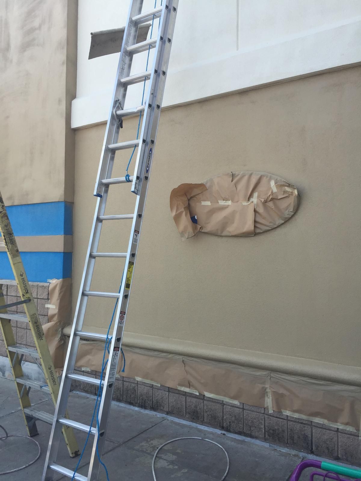 A tall ladder leans against a building being painted. Brown paper covers a circular opening.