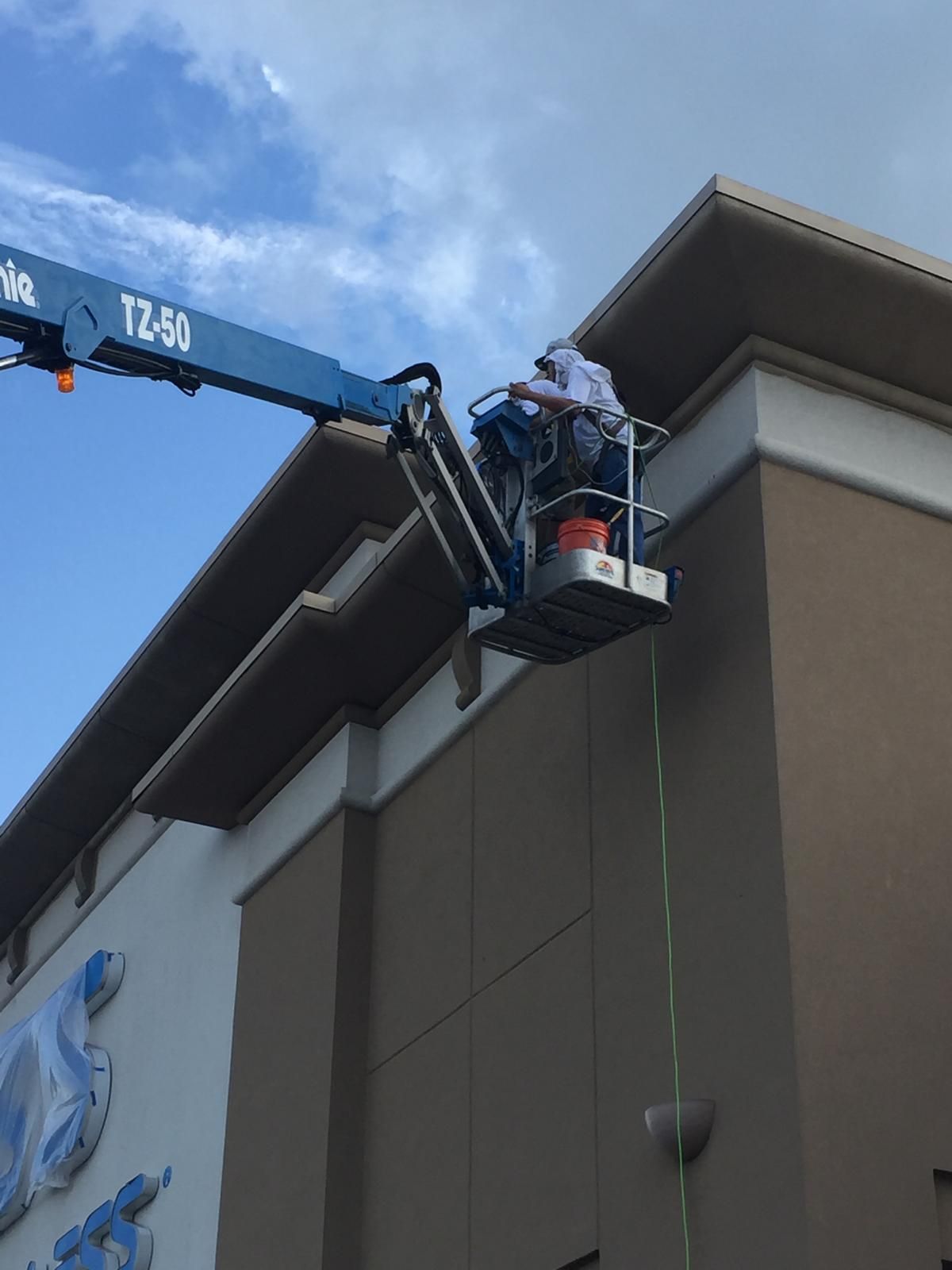 Person on a blue Genie lift, working on the edge of a building. Sky overhead, tan and white building.
