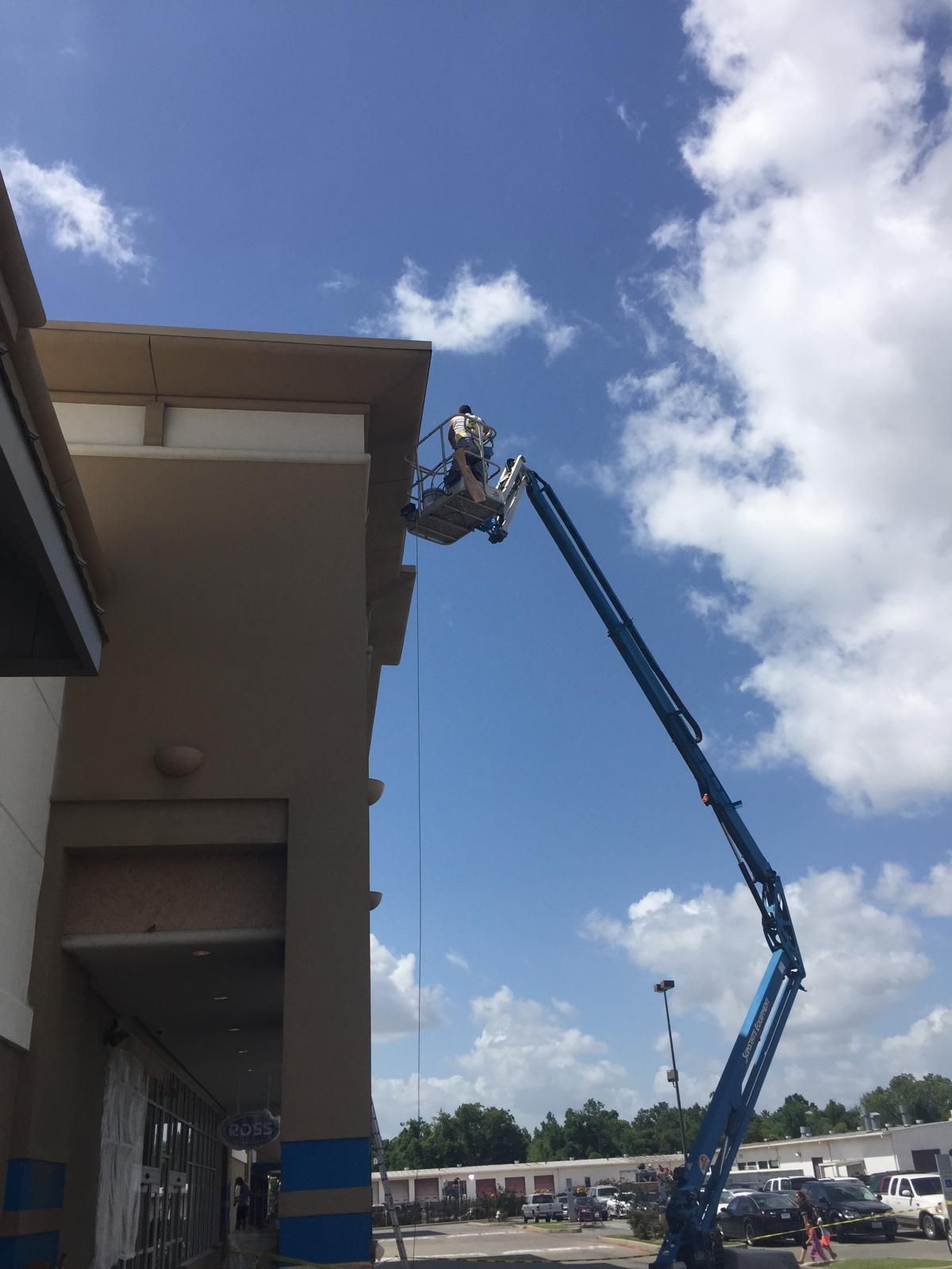 Man in lift working on the roof of a tan building under a bright blue sky.