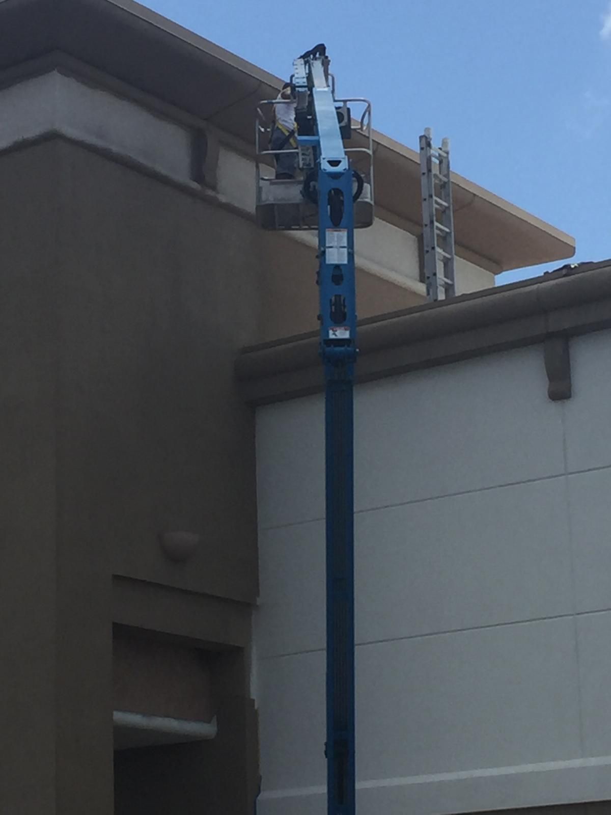 A construction worker in a blue lift painting a light-colored building against a blue sky.