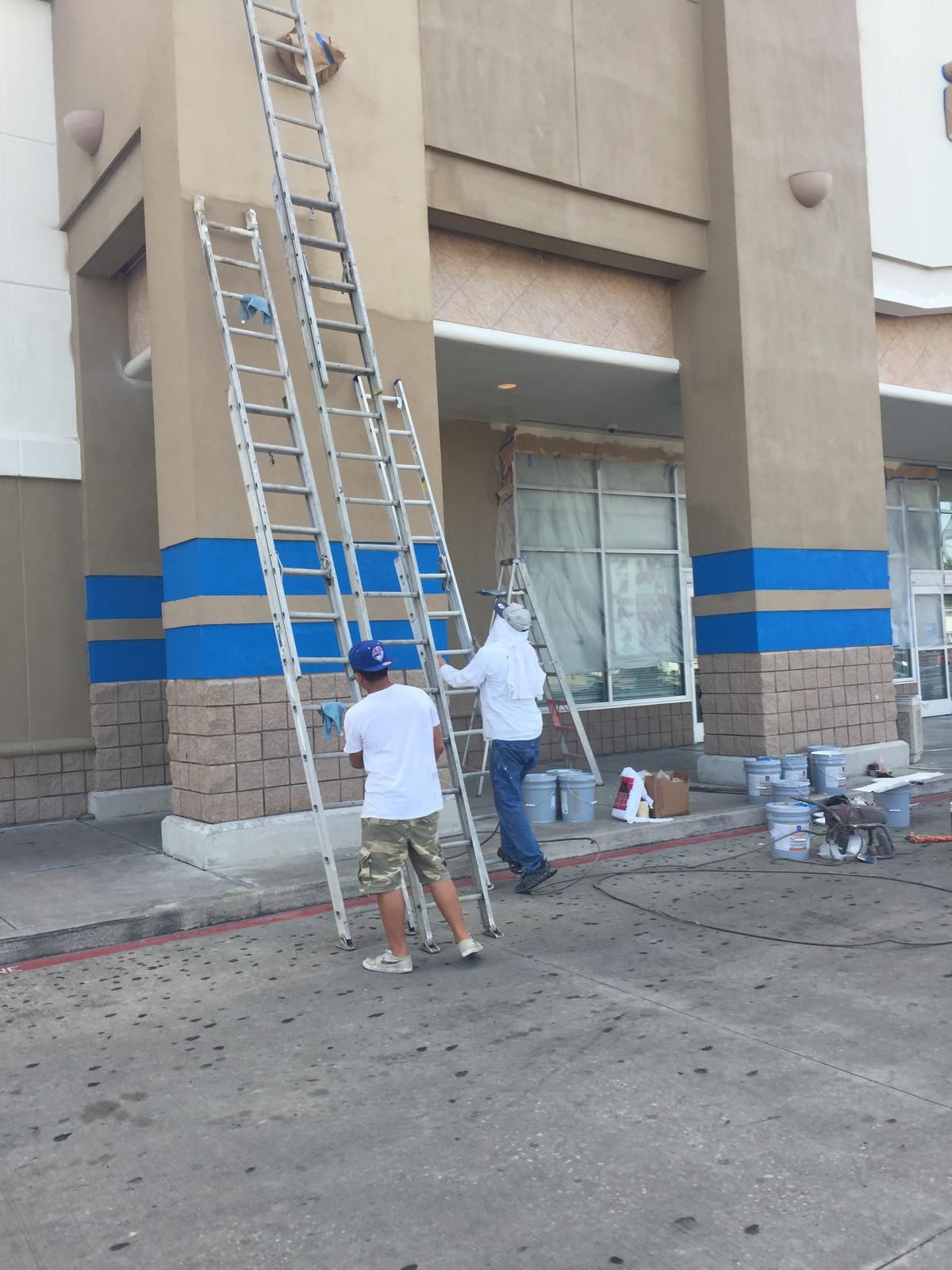 Two workers painting a building exterior. One on the ground, one on a ladder. Brown and blue color scheme.