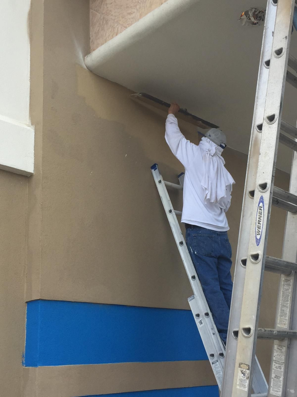 Person on a ladder painting a beige exterior wall with a blue stripe; a white cloth covers their head.