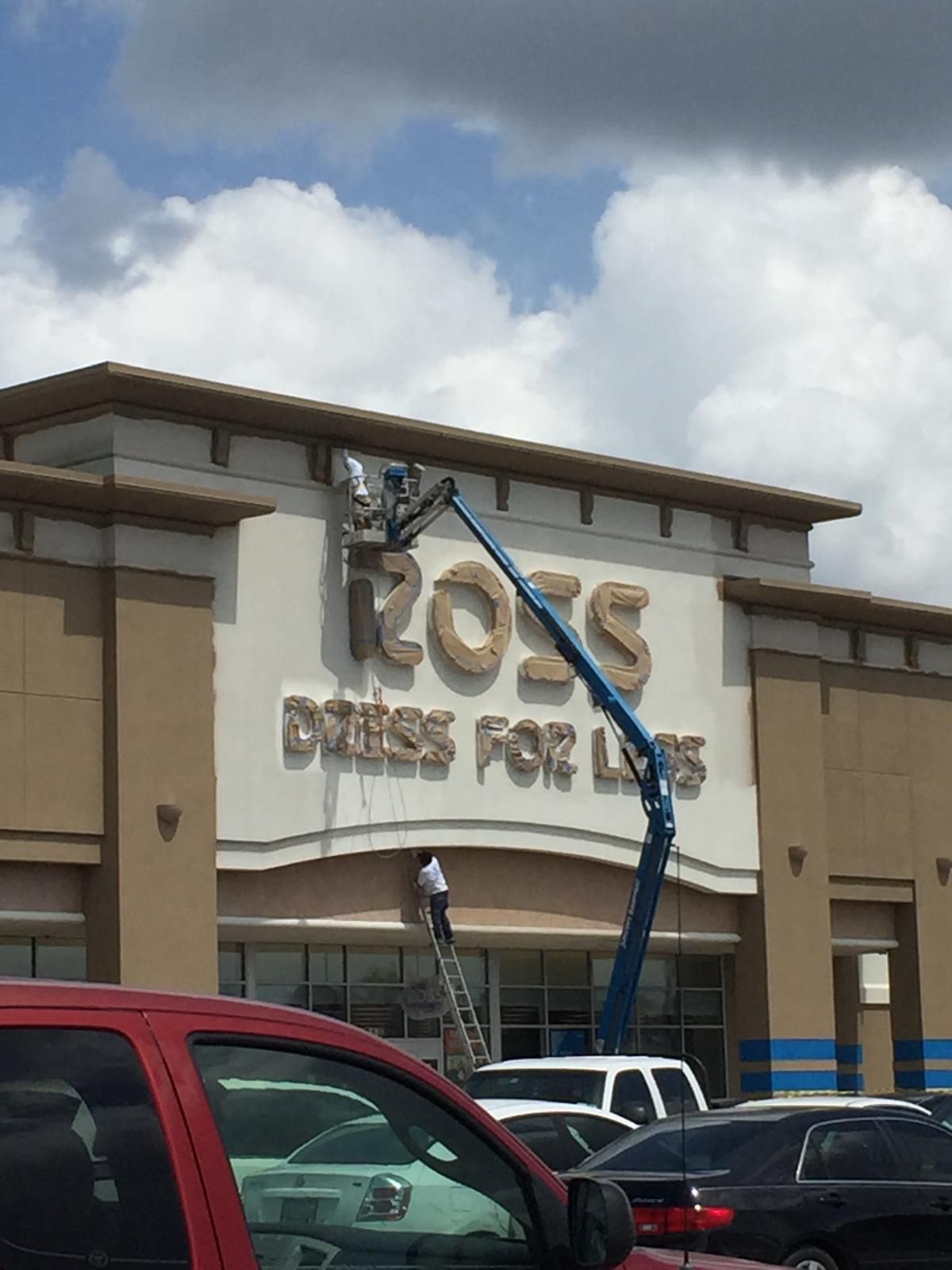 Workers on a lift installing a sign for Ross Dress for Less store in front of a cloudy sky.