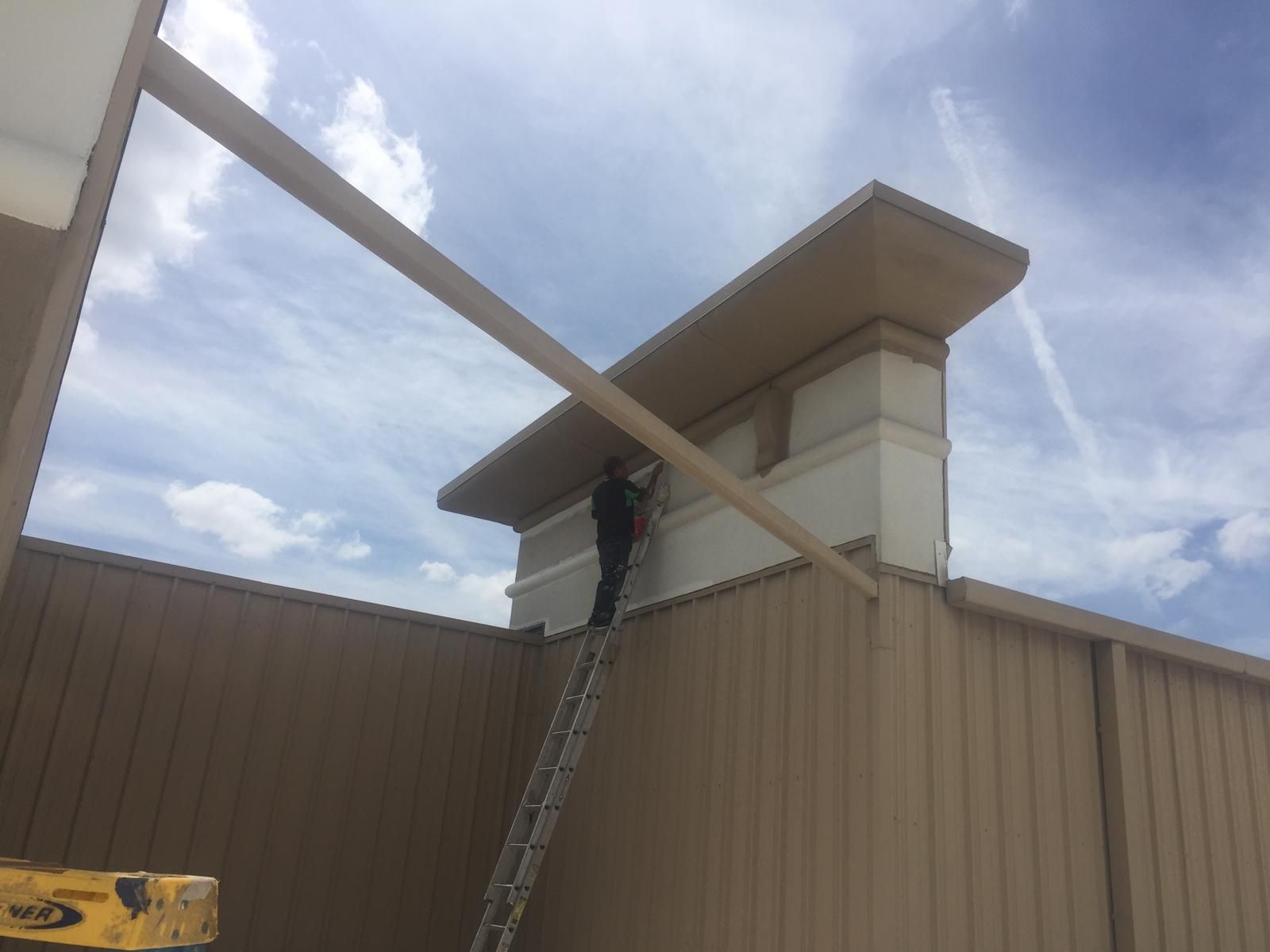 Person on a ladder installing a beam on a building's exterior, sunny sky.