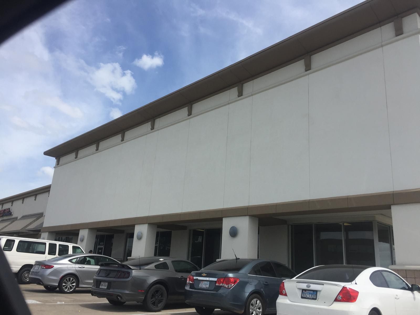 Exterior view of a strip mall with white walls and a brown awning, cars parked in front on a cloudy day.