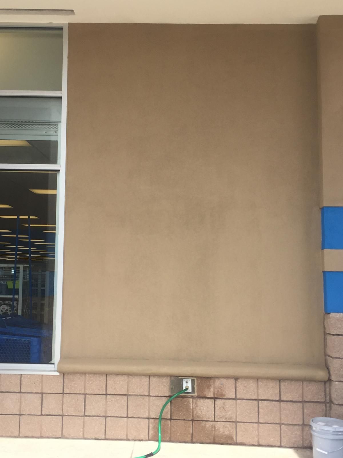 Exterior wall of a building with beige stucco, brown brick base, window, and a water hose.