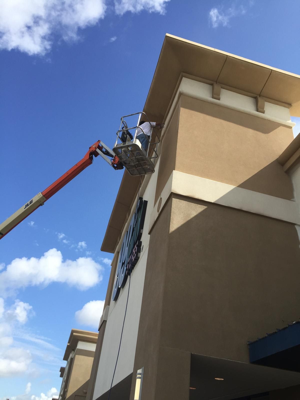 A person in a lift basket working on a sign on a tan building with a blue sky background.