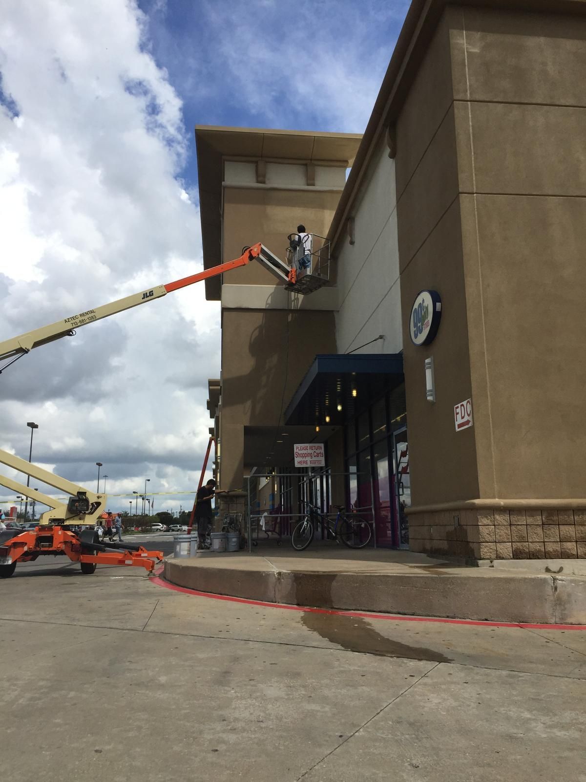 Person in lift spraying building exterior. Tan walls, blue awning, cloudy sky.
