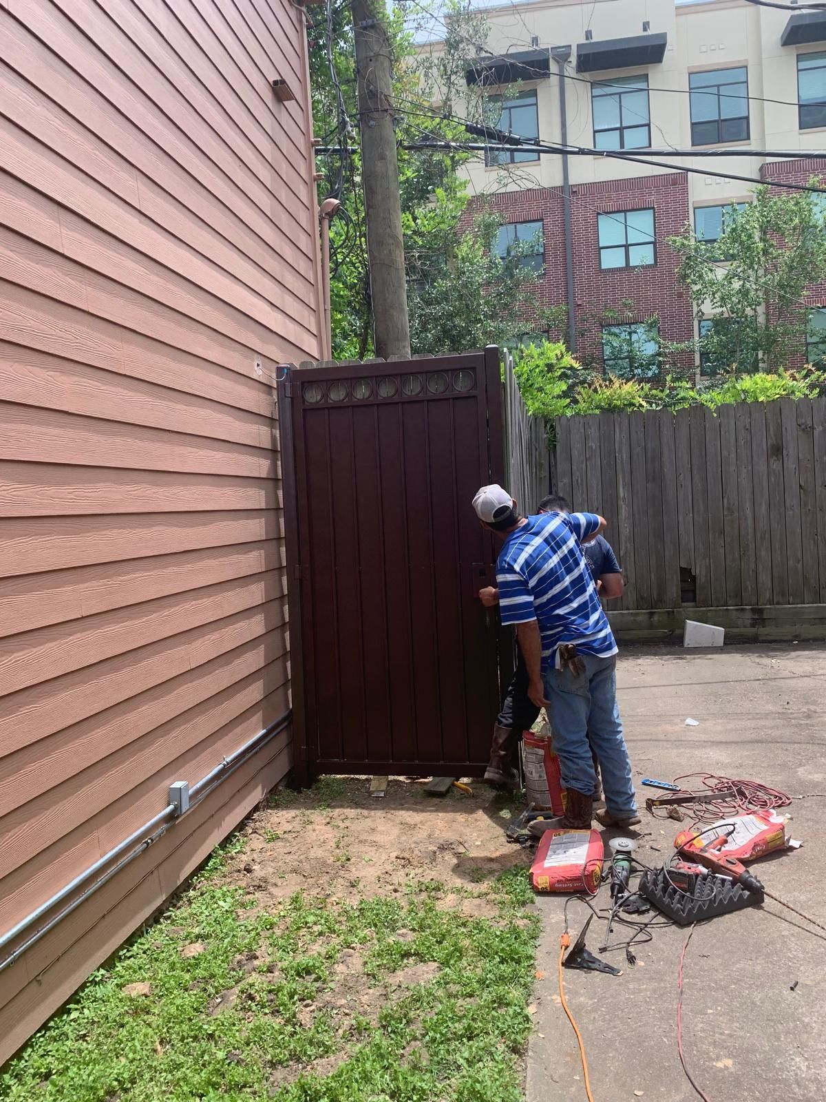Two people working on a maroon gate in a backyard, next to a brown building and a wooden fence.