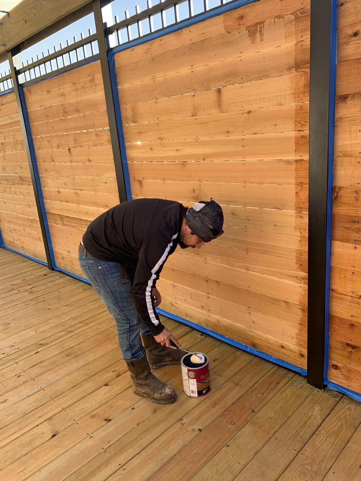 Man staining a wooden fence outdoors, using a brush and paint can.