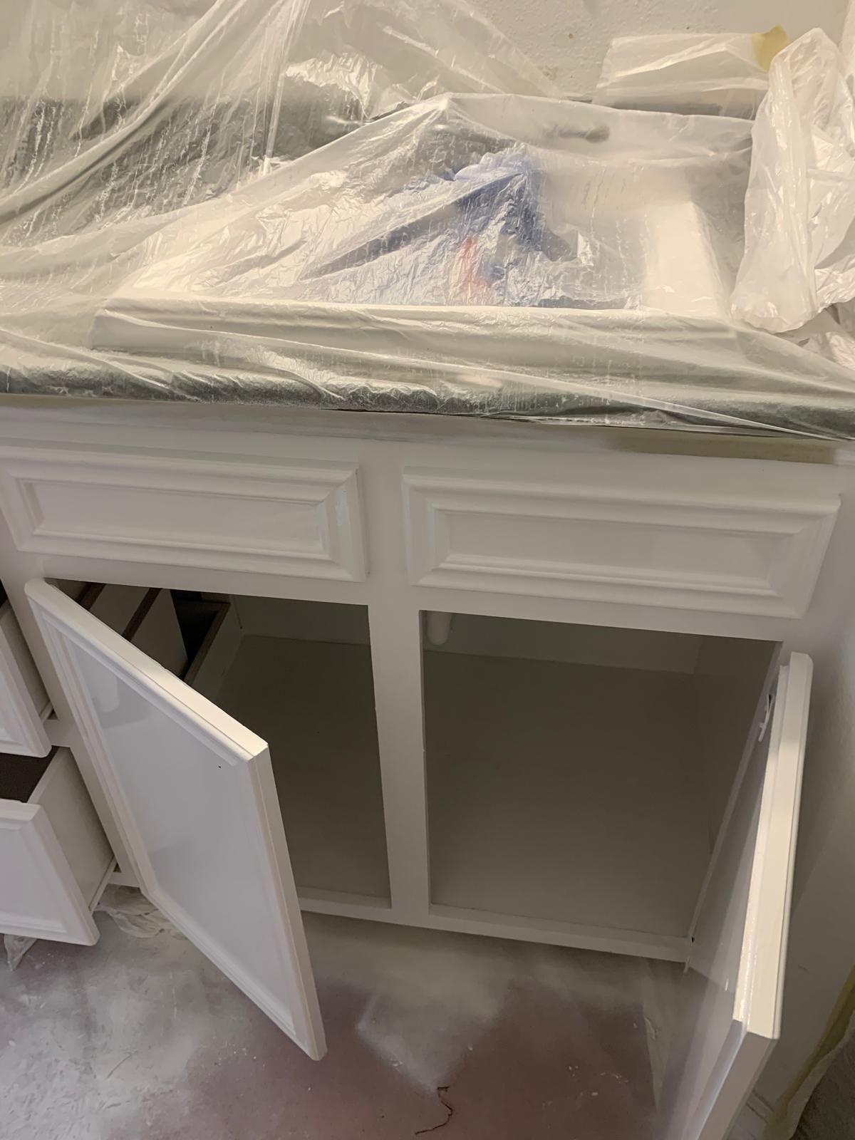 White bathroom vanity with open doors, covered in protective plastic, awaiting paint.