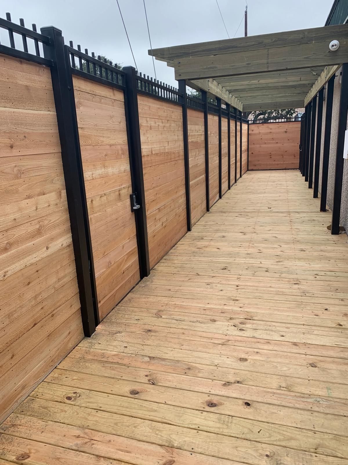 Wooden walkway between high, wood-planked fences with black posts. Overhead, a pergola offers shade.