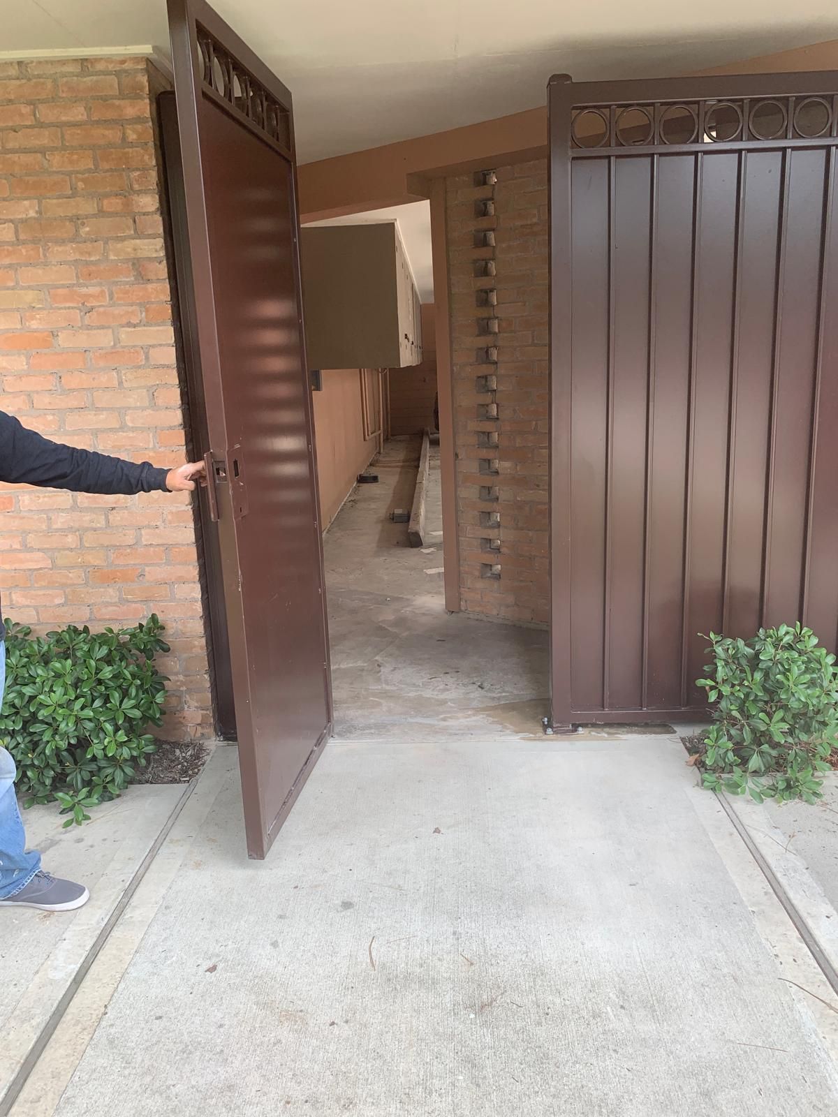 Person opening a brown metal gate leading to a walkway with brick and concrete.