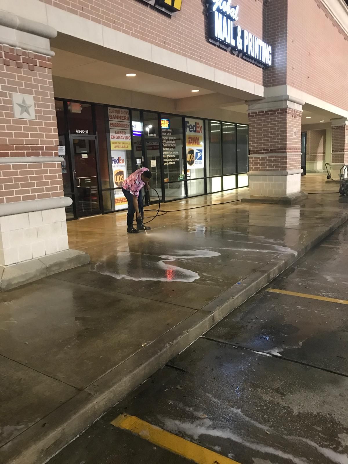 Person washing a sidewalk outside a store at night. Wet concrete, soapy water, dark background.