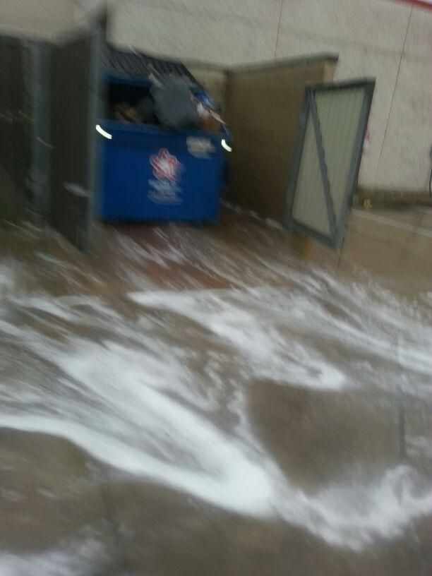 Blue dumpster with opened lid and surrounding foamy liquid on concrete.