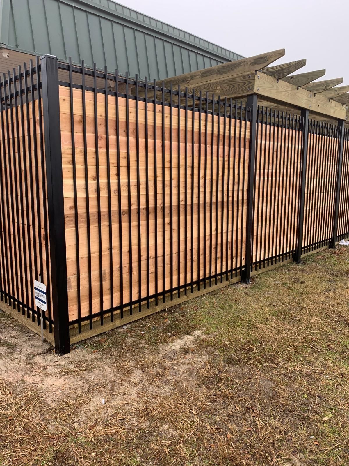 Black metal and wood fence with a pergola, in front of a green building.