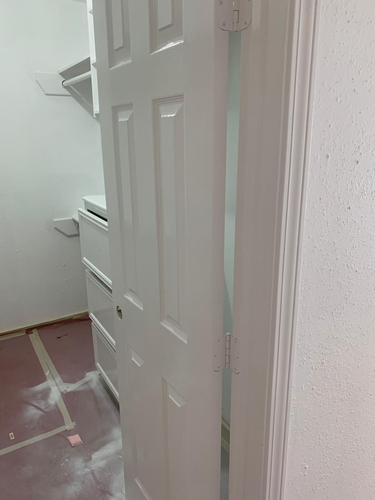 White closet door ajar, revealing white shelving and drawers inside a room. Red flooring is visible.